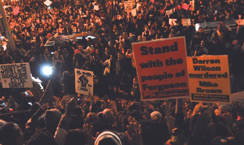 An image of a large crowd of people, some holding signs that read “Stand with the people of Ferguson” and “Darren Wilson murdered Mike Brown”.