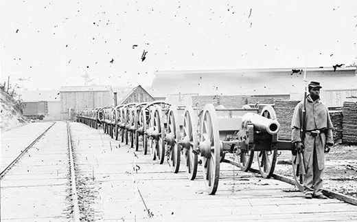 A photograph shows an African American soldier standing in front of a long line of cannons alongside a railroad track.