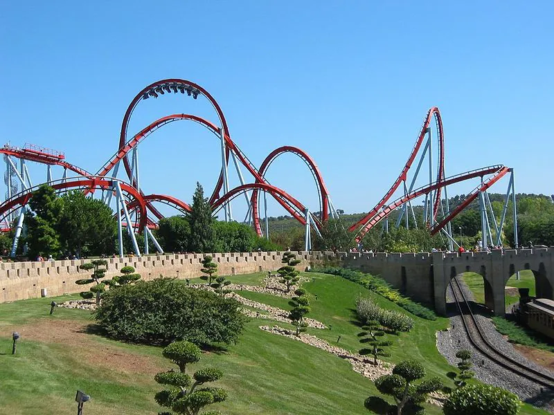 In this figure the Dragon Khan rollercoaster in Spain's Universal Port Aventura Amusement Park is shown. There are mostly curved paths in the rollercoaster. Near to the rollercoaster there is the track of rollercoaster cart under a bridge. There are some trees near the track.