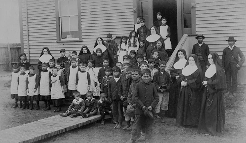 A group of White nuns and Indigenous children pose on and around steps in front of a building.