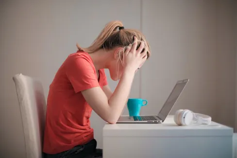 W kadrze pokazana jest z profilu młoda kobieta w czerwonej koszulce, która siedzi przed komputerem, trzymając głowę w dłoniach. 