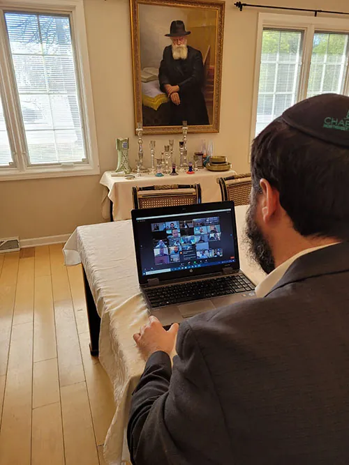 A person sits at a table with a computer. The computer screen shows a gallery view of an online meeting including many people.