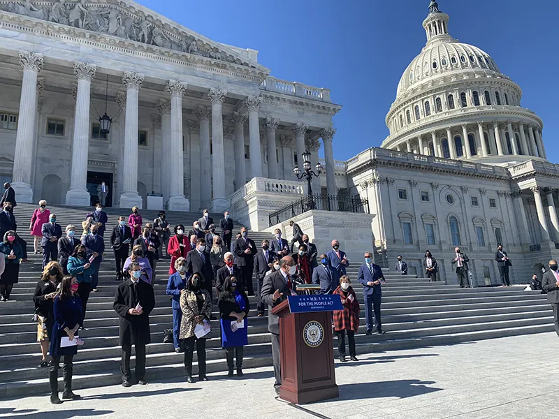 A person stands at a lectern, speaking at the foot of the steps of the United States Capitol. A group of physically-distanced people stands on the steps behind the speaker.