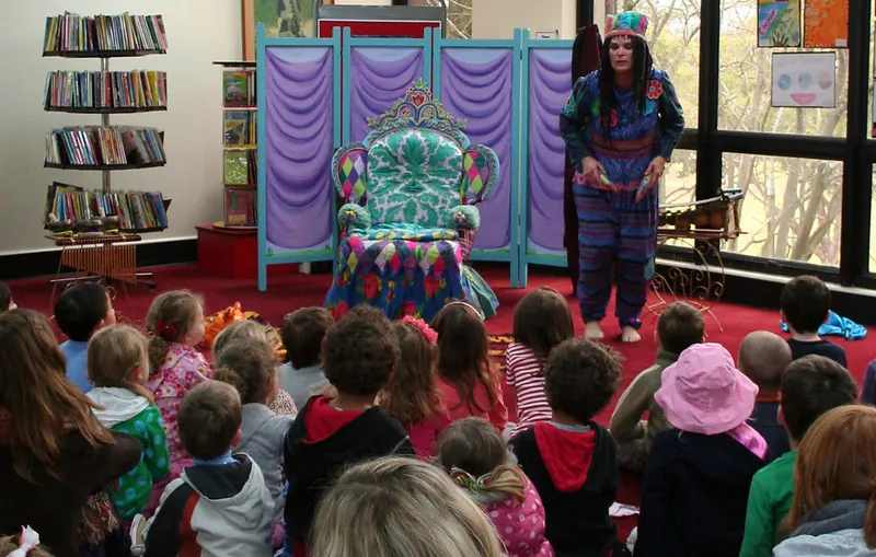 A storyteller named Bronwyn Vaughan wears a crown and tells a narrative story to the children sitting in the audience. A decorated chair and a bookshelf are pictured.
