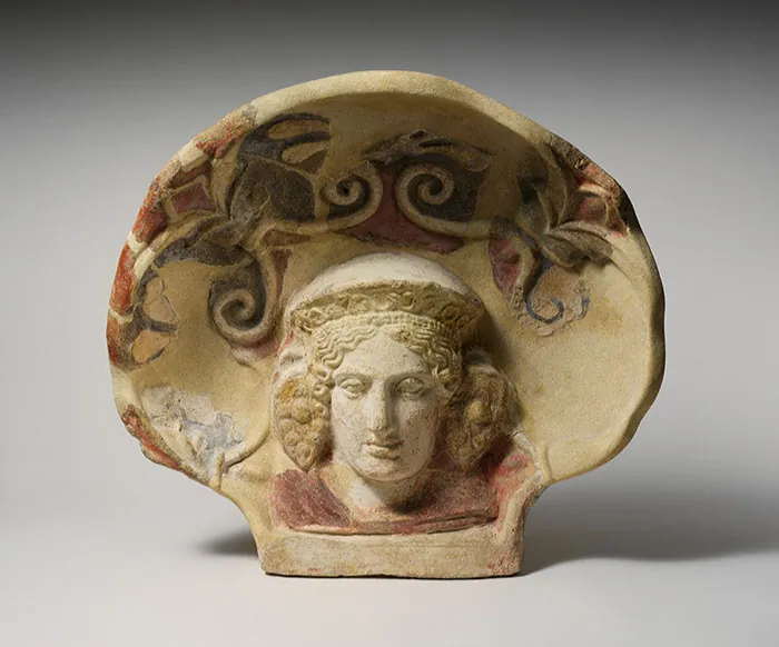 An image of a beige stoneware shell sitting upright is shown with red, black, and orange colors inside, with raised decorations. Inside the middle bottom of the shell is a carving of a head with almond shaped eyes, a large nose, and full lips. Wavy hair is seen at the forehead under a domed hat with etchings on the front. Large earrings depicted as a bunch of grapes show at the sides of the head. A reddish collar is seen around the neck. The background is light blue and the shell casts a shadow to the right.