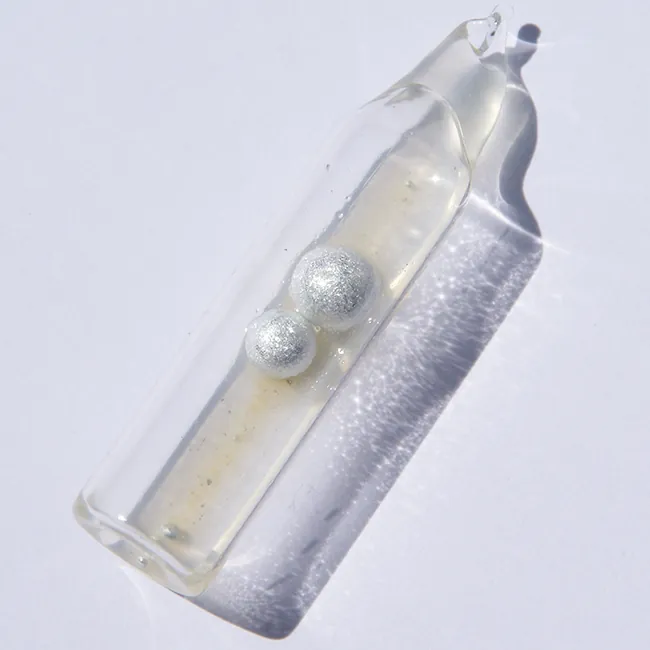 Se muestra un recipiente de vidrio sellado en forma de tubo. El recipiente está parcialmente lleno de un líquido incoloro y contiene dos esferas metálicas.
