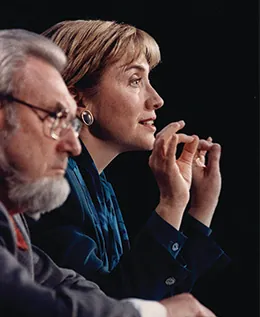 A photograph shows C. Everett Koop and Hillary Clinton in profile. They sit beside one another; Clinton speaks and gestures with her hands.