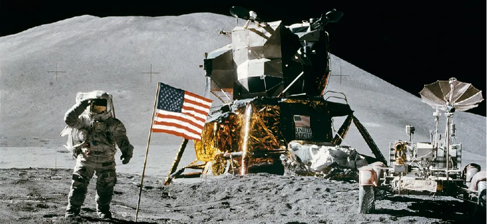Photograph of Astronauts on the Moon. At center is the landing module, and to the right is the Lunar rover used by the Astronauts to travel large distances from the landing site. At left an Astronaut salutes the American flag placed near the lander. Scattered throughout the foreground are footprints in the Lunar soil.