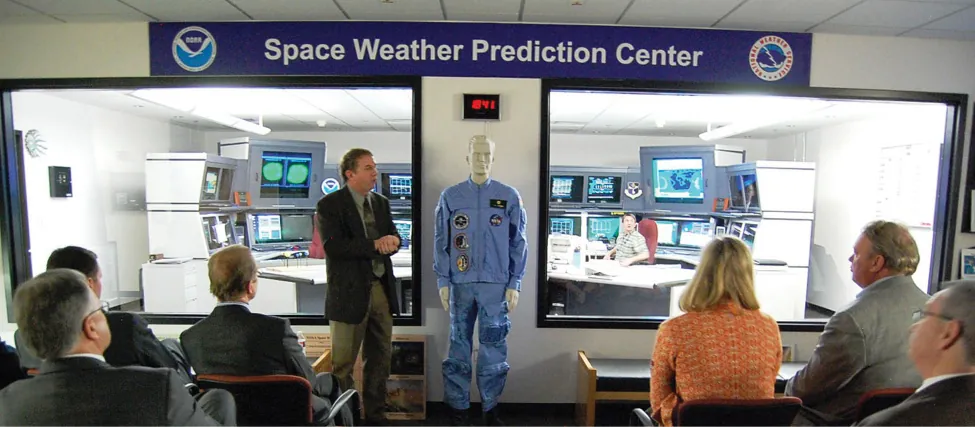 An image of several seated people in a room with a banner reading “Space weather prediction Center.” At the front of the room a person stands and addresses the seated people.