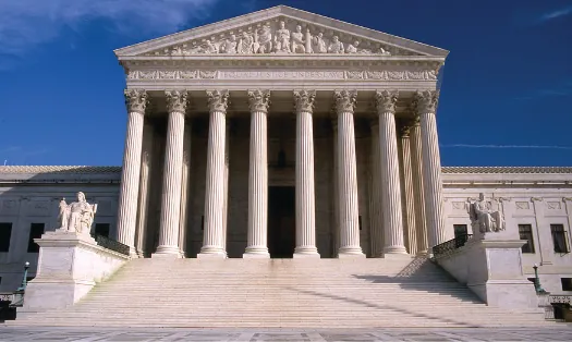 An image of the Supreme Court building. In the foreground, a set of stairs is bracketed by statues on either side, leading up to a portico. The portico has a roof supported by several tall columns.