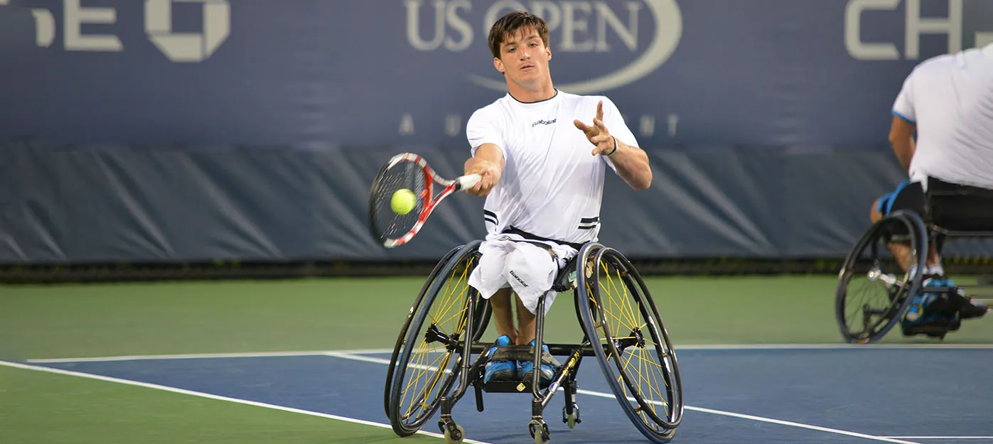 A person in a wheelchair hits a tennis ball with a racket while on a tennis court. The U.S Open logo is in the  background.