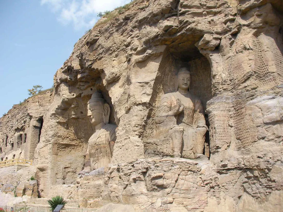 A side of a rocky mountain is seen with blue skies and sparse clouds in the left background. The left side of the hill shows square and rectangular openings and a brown fence about halfway up. On the right, two large figures are carved into the sides of the mountain. The one on the left is a head and torso. The figure wears a domed hat, has large ears with elongated lobes, and wears robes. The figure on the right is seated on a throne, smiling, with a domed hat, almond shaped eyes, and long robes and shin armor with shoes. They hold their right hand up in front of their chest. A small green bush and placard are seen in the right bottom forefront of the image as well as a short fence.