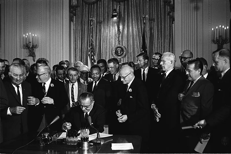 President Johnson, surrounded by onlookers, signs the landmark Civil Rights Act of 1964 . Dr. Martin Luther King Jr. stands behind the president.