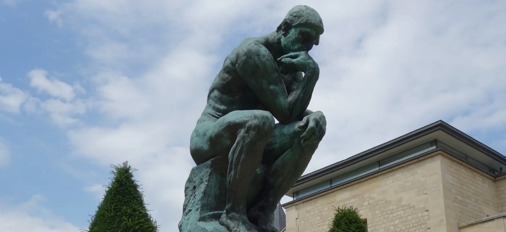 Large, bronze sculpture of a man seated in a hunched-over position, elbow on knee and chin in hand. He stares pensively into a space in the near distance.
