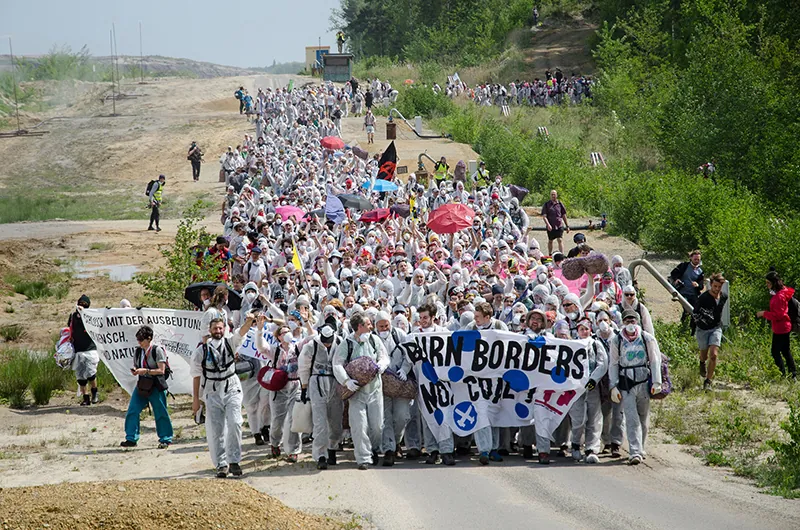 A large group of people march down a paved road surrounded by dirt, trees, and brush. Most are dressed in white. The people who lead the marchers hold a banner that says Burn Borders Not Coal.