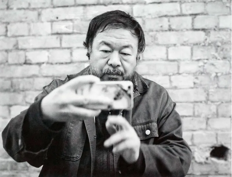 In this black and white photograph, Ai Weiwei stands in front of a brick wall looking at the screen of a cell phone he holds horizontally at the end of his outstretched arm.