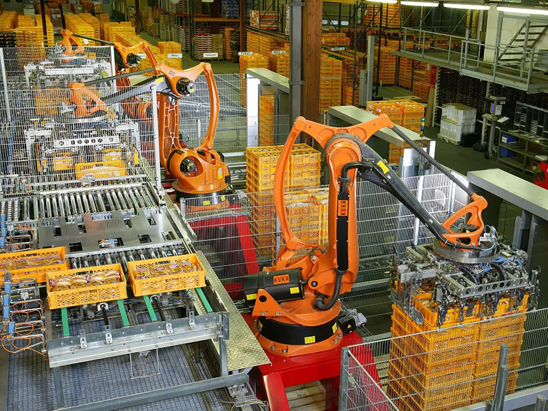 Two large robot arms with specialized devices on the ends grab and stack pallets of bread in a large factory.