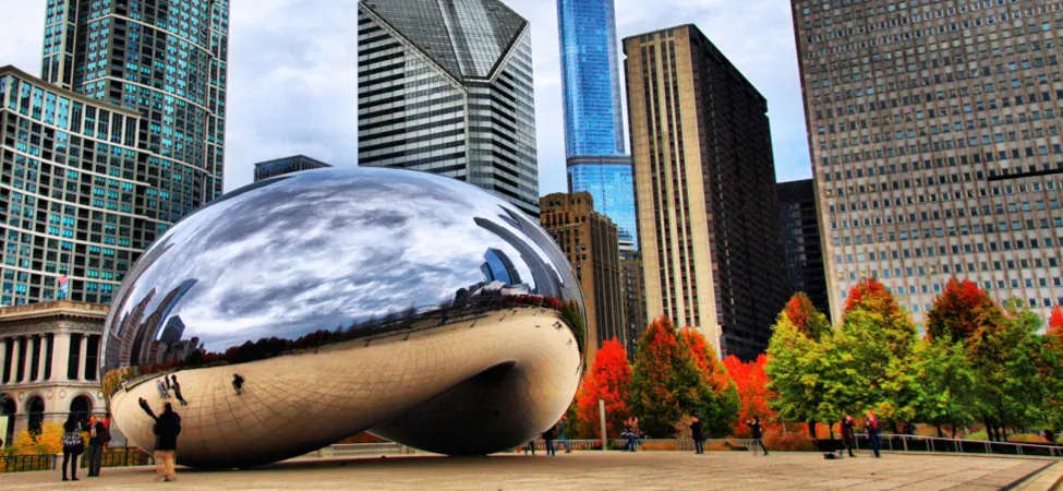 Photo shows a gigantic bean shaped structure in a plaza that is lined with trees and tall buildings. The structure’s concave underside is tall enough for people to walk under. The shiny surface of the structure reflects and distorts the image of a cloudy sky, the floor of the plaza, and the buildings and trees surrounding the structure.
