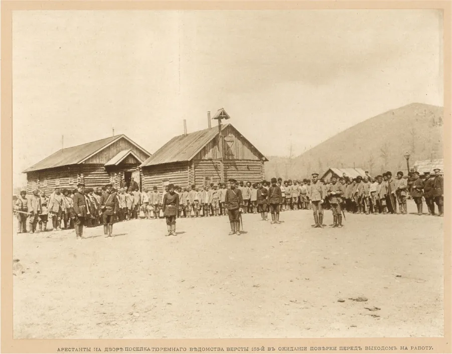 This photograph shows a group of workers standing in a line. Men in military style uniforms stand near the workers. Several log cabins are visible behind the men. A mountain is visible in the distance.