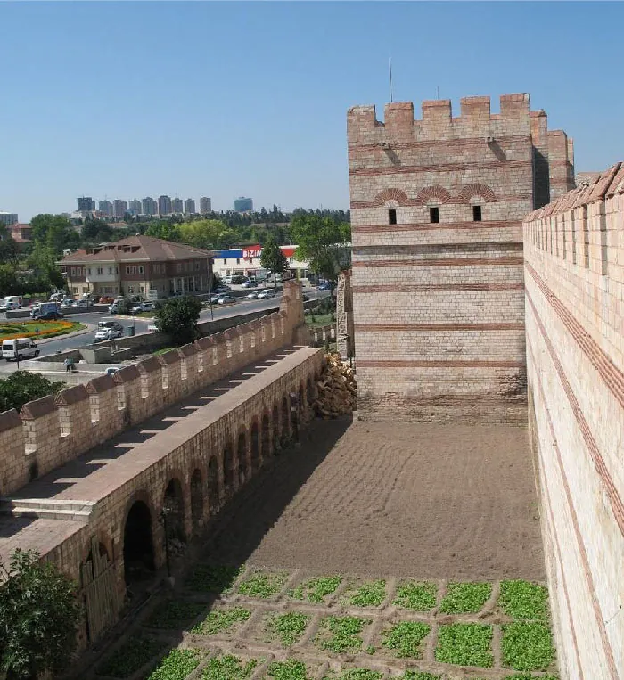A picture of a brown bricked wall with darker brown bricks making stripes is shown on the right of the image. In front of the wall is an area filled with dirt at one end and squares of green vegetation at the forefront. At the end of the dirt stands a brick tower with three openings toward the top and notches across the top. To the left of the dirt area is another wall with arched openings on the right side and notches at the top on the left side of the wall. In the far left background a short brown building can be seen with two levels of rectangle windows and a pointed roof. A parking lot filled with cars and another white, red, and blue building can be seen. Beyond the brown building green trees are seen and ten tall gray rectangular buildings stand in the very far background. A clear blue sky shows.