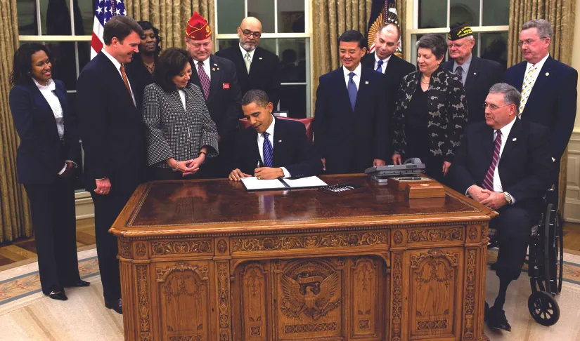 An image of a group of 12 people standing around Barack Obama, who is seated at a desk and signing a piece of paper.