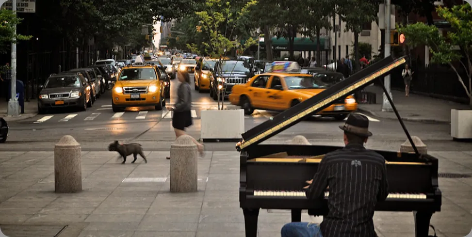 A photograph shows a person playing a piano on the sidewalk near a busy intersection in a city. Zdjęcie ukazuje osobę grającą na pianinie na chodniku u zbiegu ruchliwegi skrzyżowania.