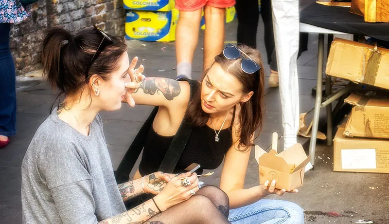A photo shows two young women sitting on the edge of a busy sidewalk eating a meal. One reaches to the other’s face as if to remove a piece of food.