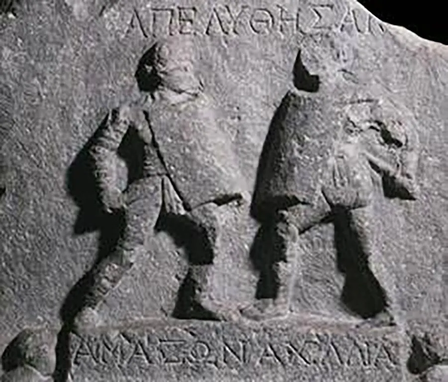 A carving on a gray stone is shown on a black background. The image is worn and lacking details. Two figures face each other with curved rectangular shields at their fronts and weapons in their hands. No facial features are shown. The letters “AnEAYOHEA” are carved above and the letters “AMAZONAXANA” are carved below the figures.