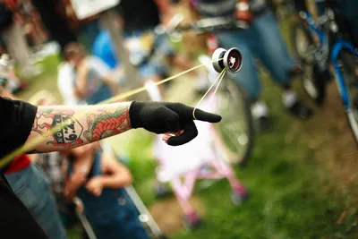 The figure shows the left arm of a man with tattoo imprints and wearing a glove. He is circulating a yo-yo toy, which is in mid air and connected by the string to his hand. Some people are standing in the background watching the yo-yo trick.
