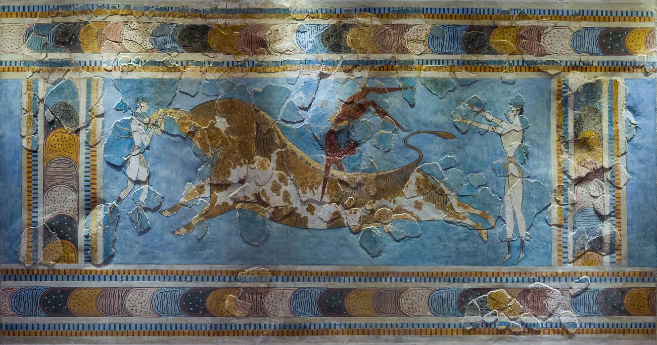 An image of a rectangular wall painting is shown. The surface of the wall is stone with flat, round areas protruding out in various locations. The borders are beige, brown, and blue stripes with crescent shapes, with dots, lines and dashes inside the crescents. In the middle of the frame, a large brown and white animal with short legs and horns is stretched across the blue background with its legs splayed out. A dark brown figure in a light brown loincloth is upside down on the middle of the animal with their legs kicking to the right. A figure stands in front and behind the animal. The one in front wears a white short robe and is grasping the animal’s front horn. The figure behind the animal wears a brown loincloth, wrist and arm bangles, and is holding both arms straight out toward the brown figure on the animal’s back.