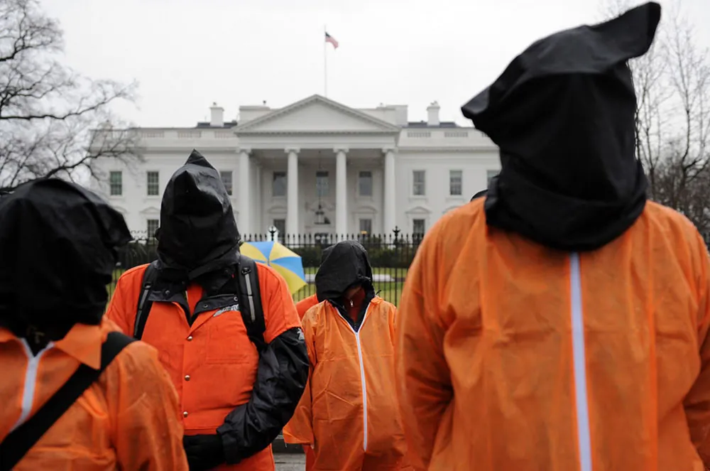 Several people stand in front of the White House wearing prison jumpsuits with hoods over their heads.