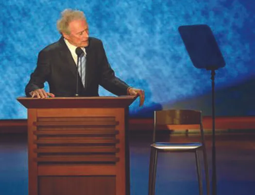 An image of Clint Eastwood standing behind a podium. Next to him on the right is an empty chair.