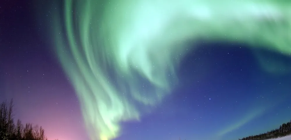 A shimmering curtain of green lights in the sky above a snow covered landscape. Stars are visible in the dusky sky beyond the lights.