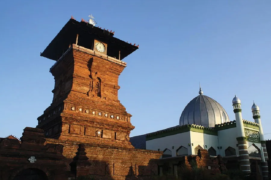 This photograph shows the Menara Kudus Mosque complex. To the left is a brown tower with a clock at the top. To the right is a building topped with a large silver dome and two smaller sliver domes.