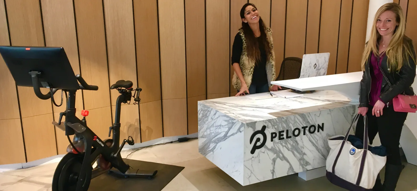 One person stands behind a marble desk with the Peloton logo on it, while another person stands in front of the desk holding a tote bag. A Peloton bike is to the right of the desk.