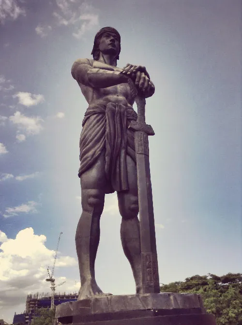 This photograph shows a bronze statue of Lapulapu who wears a loincloth and holds a large sword.