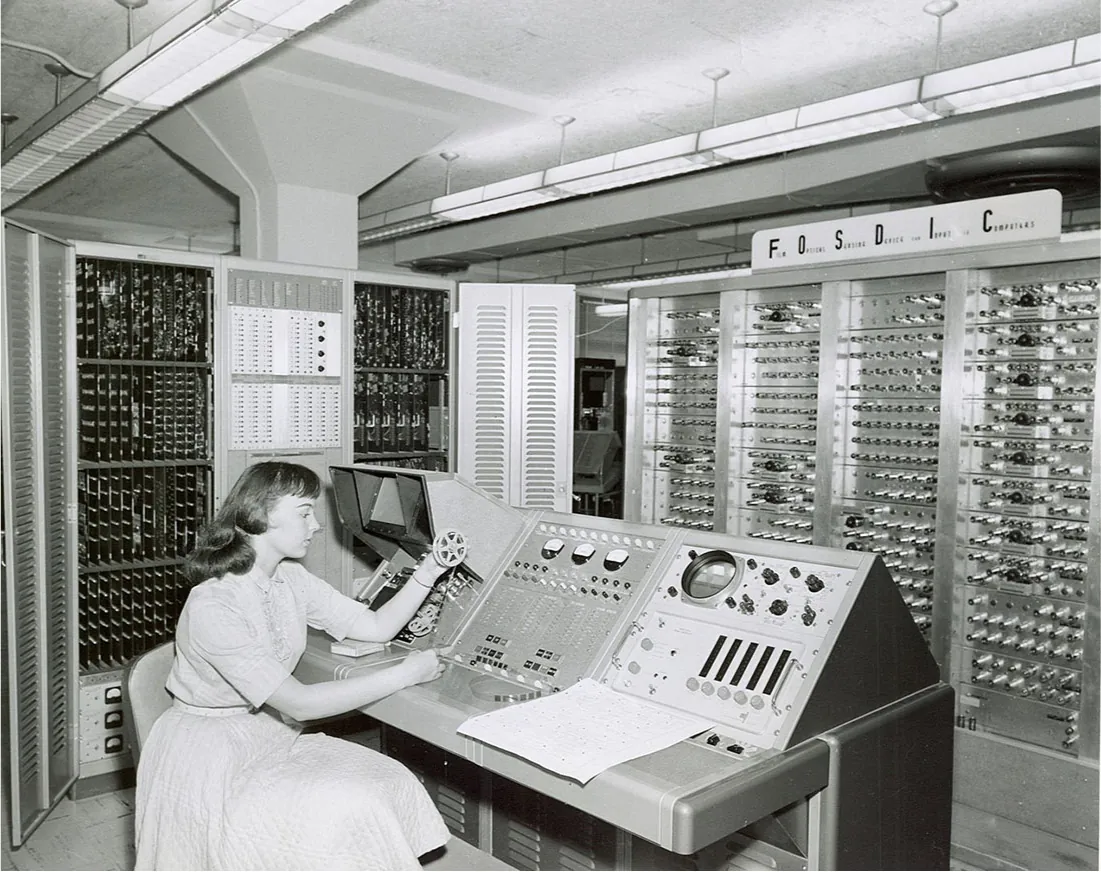 A black and white photo is shown of a woman in a light-colored belted dress with short sleeves sitting at a table with a large machine on it. She has long hair and bangs and is holding a spool of film in her left hand, pulling the film down with her right hand. There are many buttons, knobs, and dials on the machine in front of her and vents underneath the machine by her knees. In the background the walls around her hold machinery with knobs, slats, and buttons all in rows on several panels. On the right wall along the top is a sign with “Film Optical Sensing Device for Input to Computers” printed in black across it. Long, thin, slatted lights hang from the ceiling.