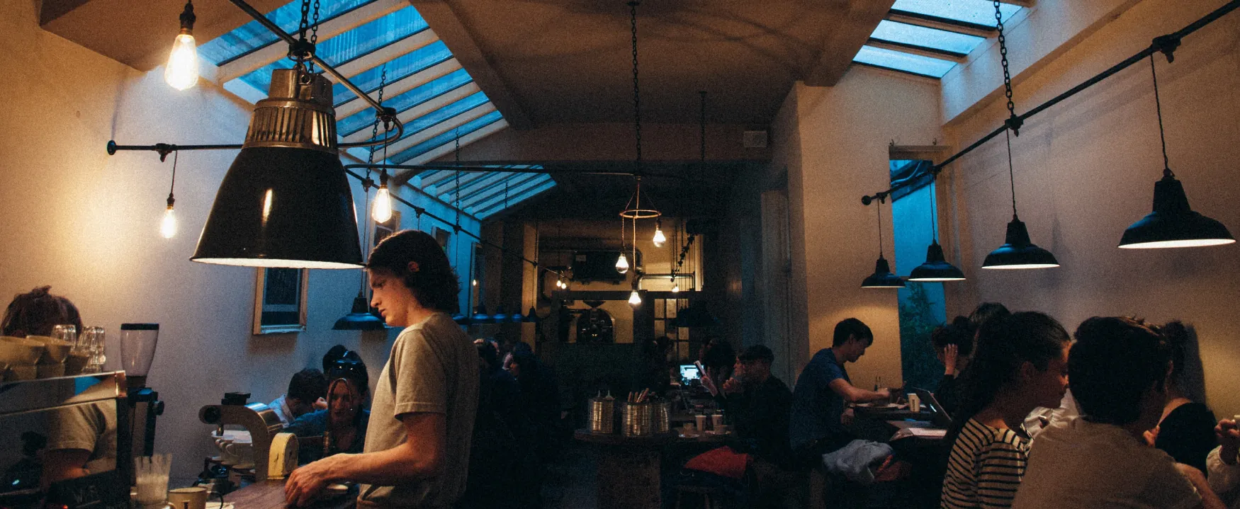 A dimly lit, crowded coffee shop is shown.  Patrons sit at tables, and a worker attends to a customer at the counter.