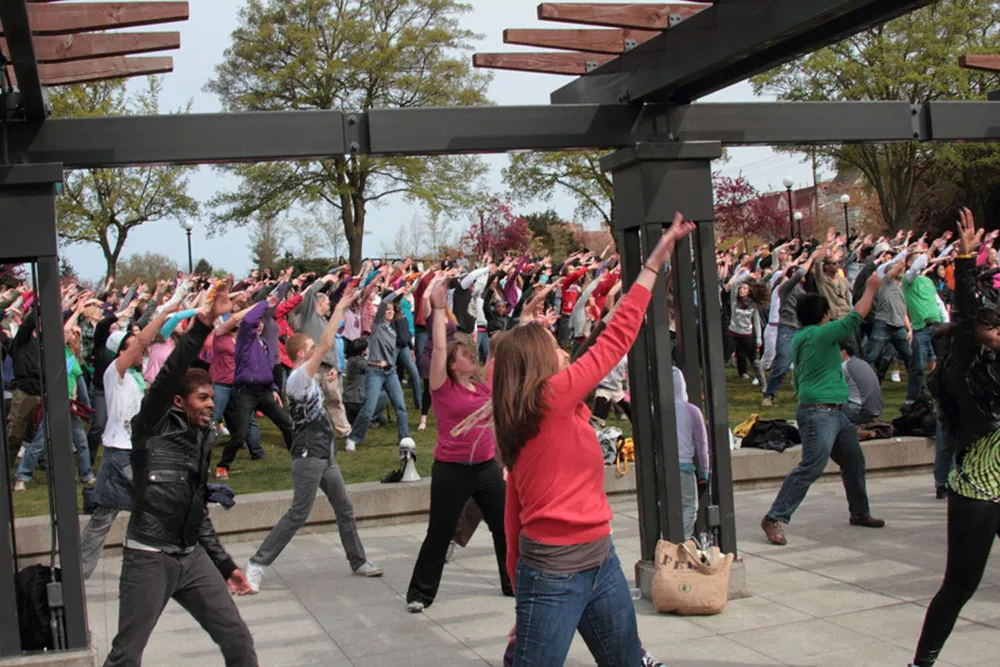 A large group of people in casual and diverse clothing make identical movements in what appears to be a park or plaza.