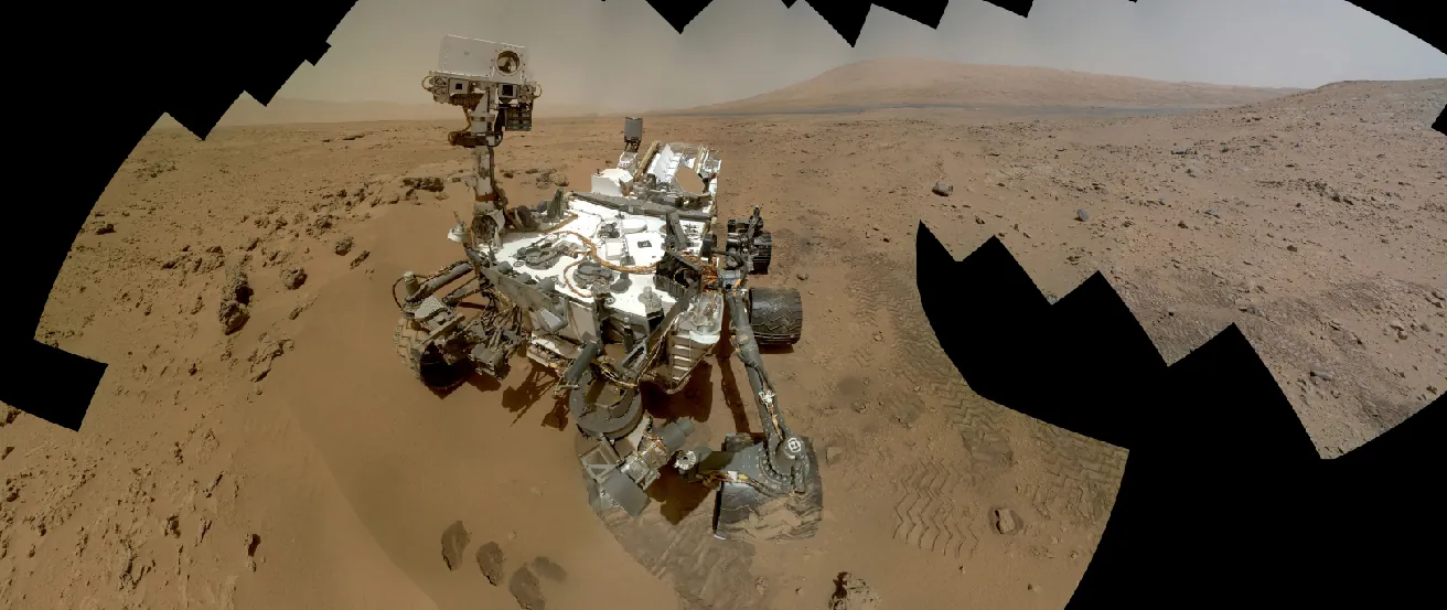 Image of the Curiosity Rover on the Martain surface. In this composite photograph we see the rover perched on the rusty-red Martian soil, with a series of hills and a dusty colored sky in the background. The outline of this image is jagged due to the effects of combining the individual frames into a single montage.