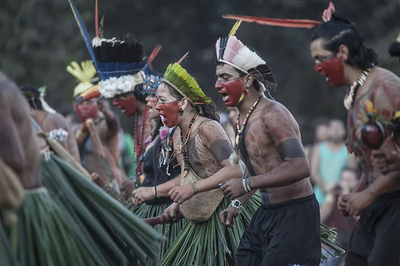 A group of people wearing colorful feather headdresses and body paint perform a group dance.