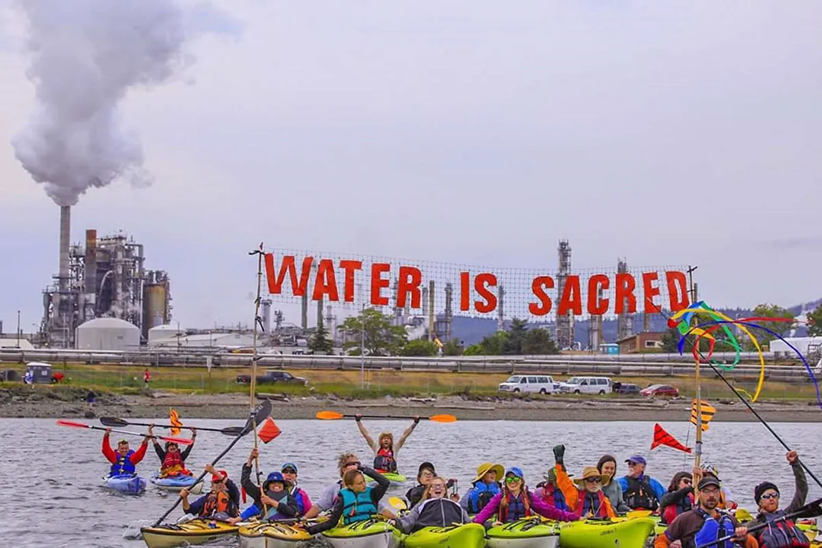 About 20 people in kayaks float in front of an oil refinery. They hold a sign that reads “Water Is Sacred.”