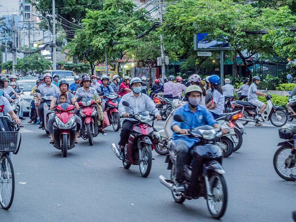 About twenty people on motorcycles and scooters drive along a crowded road with cars and bicycles. Some wear masks and most wear helmets.