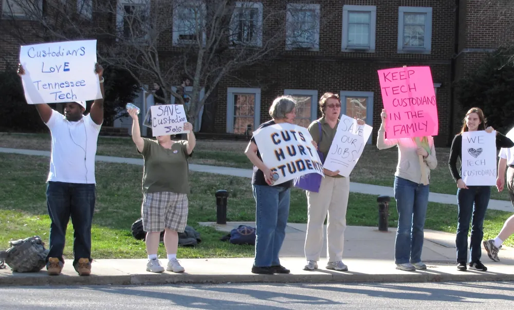 A group of people are shown standing on a sidewalk holding protest signs.