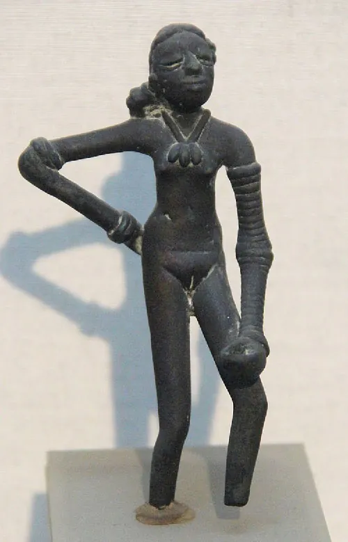 A picture of a black statue of a lady on an off-white platform is seen. Her shadow reflects behind her on a white background. The statue has long hair in a low ponytail with closed eyes and a half-smile on her face. She wears a necklace with three oval objects hanging in front. Her right hand rests on her hip and her left arm hangs down with her fist resting above her bent left knee. Bangles adorn both her arms. Her feet are missing and her right leg is anchored to the platform below while the left leg is resting in the air.