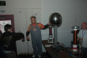 This is a photograph of a man touching the large sphere of a Van de Graaff generator, causing his hair to stand on end. The picture also shows two observers, one of whom has extended his right arm toward the man touching the generator.