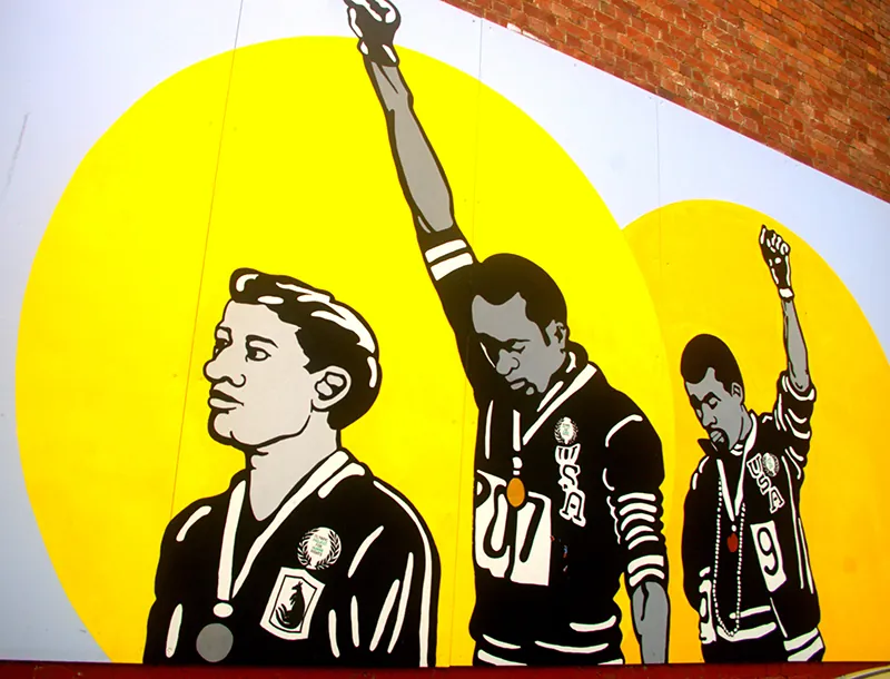 Mural on a wall depicting three athletes with medals around their necks. Two bow their heads and hold their fists up on the air.