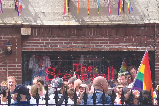 An image of a group of people standing in front of a brick building. A sign in the window of the building reads “The Stonewall”. A multicolored flag is held by a person to the right.