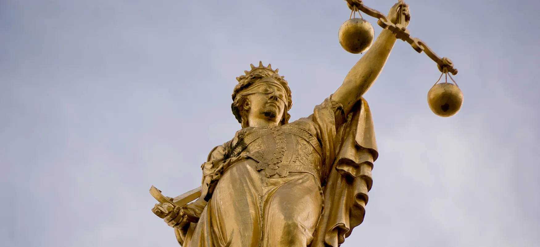 In a golden statue Lady Justice is blindfolded. In one hand she holds a balanced scale, and in the other hand she holds a sword tucked under her arm.