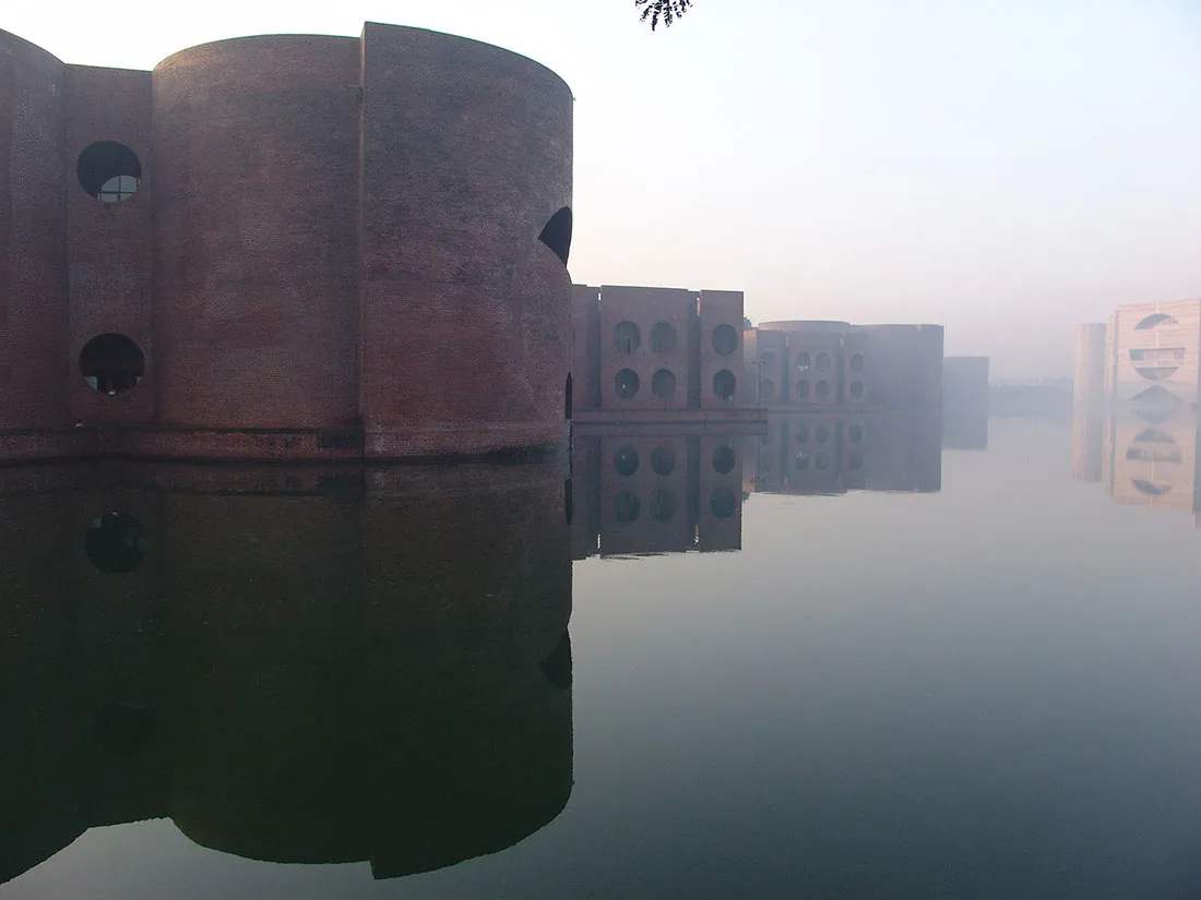 The National Assembly Building portion of the Capitol Complex in Dhaka, Bangladesh. A brick building with rounded walls and large circular open windows sits in a lake. In the background, another building in the complex with an entirely different design sits within the lake.
